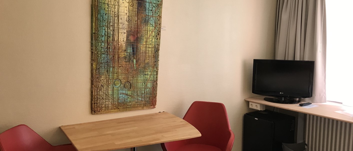 Sitzecke in einem Doppelzimmer mit zwei roten Stühlen, einem Holztisch, einem Fernseher und einem bunten Kunstwerk an der Wand., © Abalon Hotel Ideal Sitzecke in einem Doppelzimmer mit zwei roten Stühlen, einem Holztisch, einem Fernseher und einem bunten Kunstwerk an der Wand., © Abalon Hotel Ideal