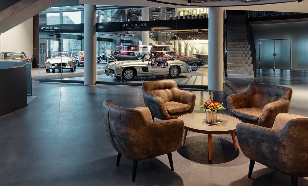 Moderne Lobby mit vier Sesseln um einen Tisch, Blumenarrangement. Im Hintergrund sind Oldtimer hinter Glas ausgestellt., © V8 HOTEL Motorworld Region Stuttgart
