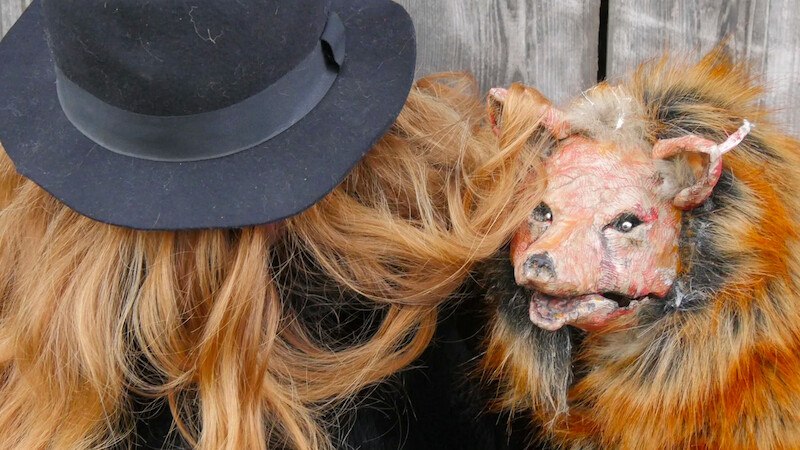 A person with long hair and a black hat standing next to a fur-covered animal mask against a wooden background., © FITZ Das Theater animierter Formen A person with long hair and a black hat standing next to a fur-covered animal mask against a wooden background., © FITZ Das Theater animierter Formen