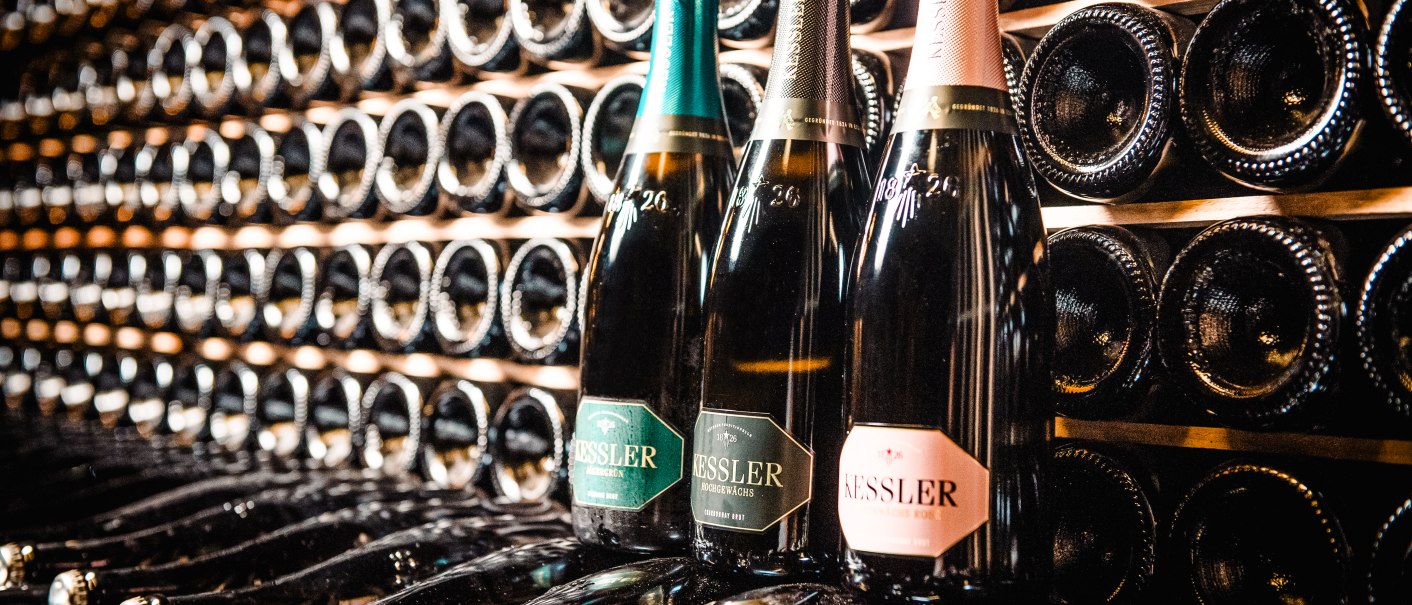 Drei Kessler Sektflaschen stehen vor einem Regal voller liegender Flaschen. Die Etiketten sind in Grün, Schwarz und Rosa gehalten., © Sarah Dulay Drei Kessler Sektflaschen stehen vor einem Regal voller liegender Flaschen. Die Etiketten sind in Grün, Schwarz und Rosa gehalten., © Sarah Dulay