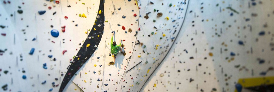 Ein Kletterer in grüner Kleidung erklimmt eine hohe Indoor-Kletterwand mit bunten Griffen im DAV Kletter- und Boulderzentrum Schwaben., © Rockerei Ein Kletterer in grüner Kleidung erklimmt eine hohe Indoor-Kletterwand mit bunten Griffen im DAV Kletter- und Boulderzentrum Schwaben., © Rockerei
