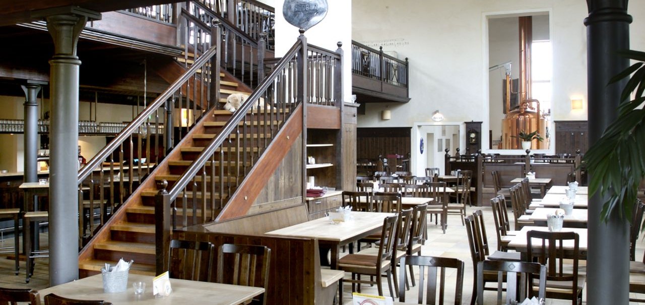 Ein Brauhaus mit Holztischen und -stühlen, einer Treppe und einem Kupferkessel im Hintergrund. Helle, geräumige Atmosphäre., © TOMAS Ein Brauhaus mit Holztischen und -stühlen, einer Treppe und einem Kupferkessel im Hintergrund. Helle, geräumige Atmosphäre., © TOMAS