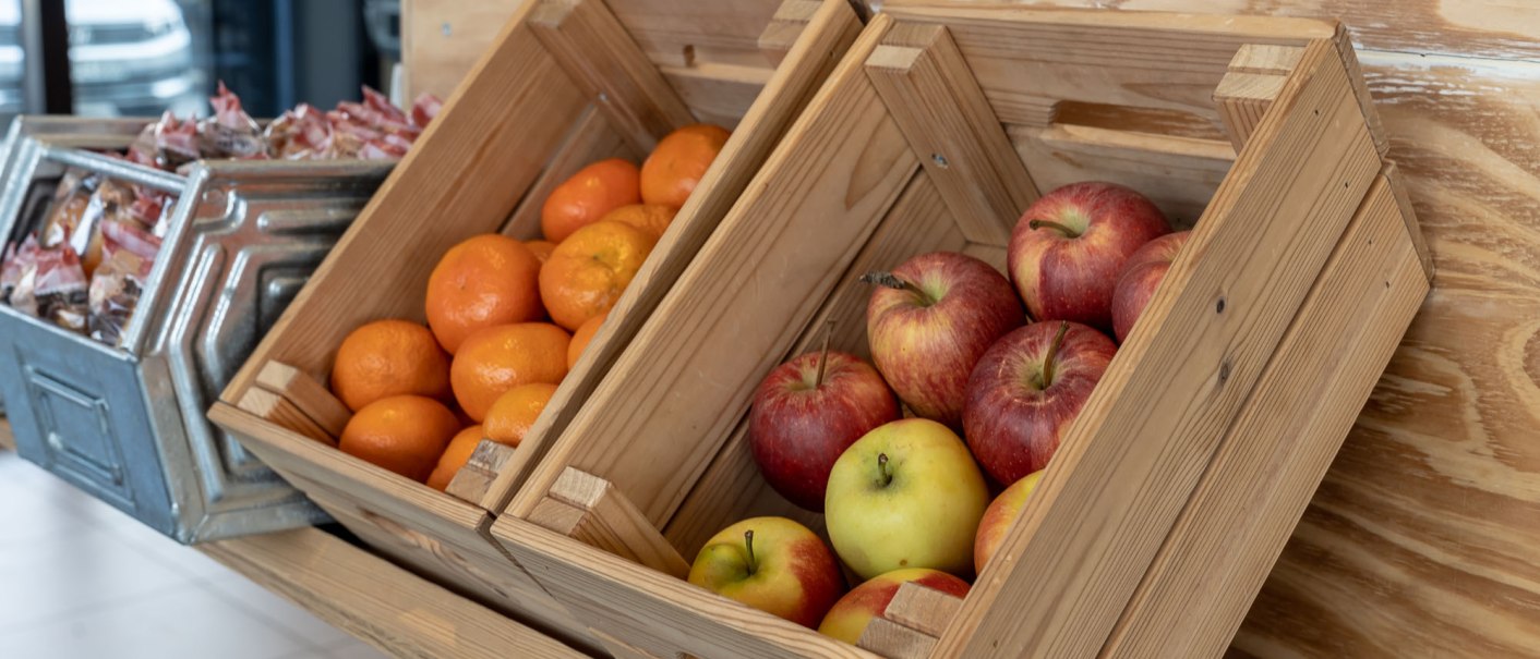 Wooden crates with fresh apples and oranges, next to a metal container with packaged snacks in a breakfast area., © a2 HOTELS