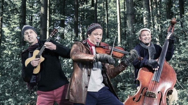 Drei Musiker stehen im Wald und spielen Instrumente: Gitarre, Geige und Kontrabass. Sie tragen warme Kleidung und wirken fröhlich., © Theaterhaus Stuttgart e.V. Drei Musiker stehen im Wald und spielen Instrumente: Gitarre, Geige und Kontrabass. Sie tragen warme Kleidung und wirken fröhlich., © Theaterhaus Stuttgart e.V.