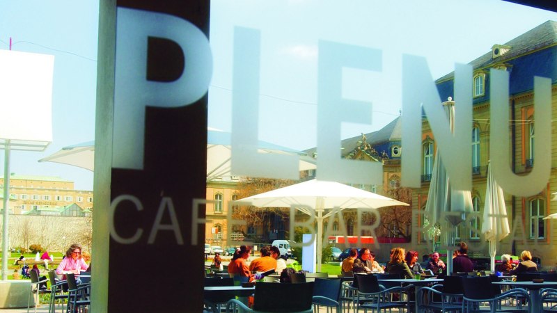 People sit under parasols in the outdoor area of a café. Historical buildings can be seen in the background., © SMG