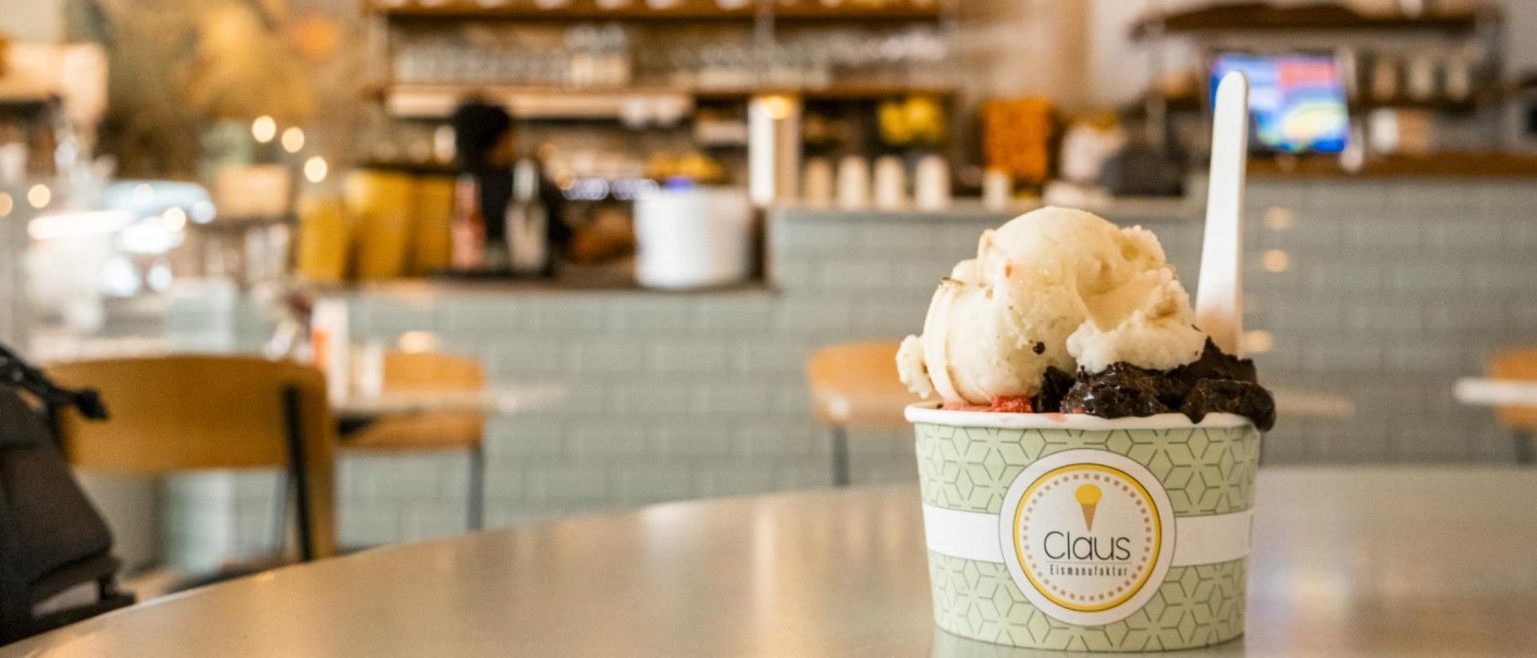 Ein Becher Eiscreme von Claus Eismanufaktur steht auf einem Tisch in einem Café. Der Hintergrund ist unscharf und zeigt eine Theke., © SMG, Sarah Schmid