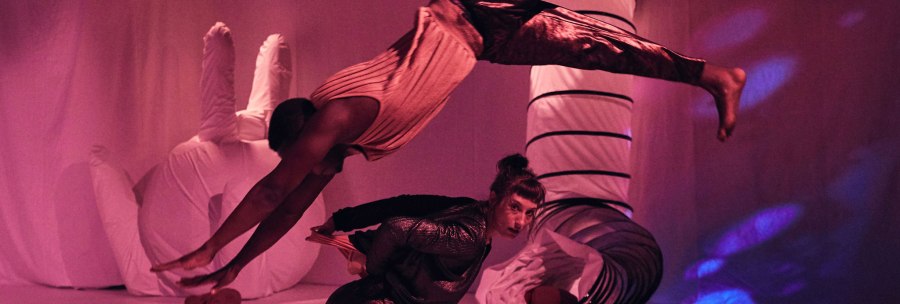 Two acrobats in a pink-lit room. One jumps elegantly over the other, who remains in a crouched position., © JES, Alex Wunsch