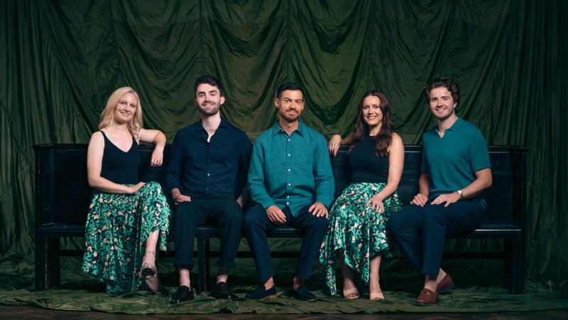 Fünf Personen sitzen lächelnd auf einer Bank vor einem grünen Vorhang. Zwei Frauen tragen gemusterte Röcke, die Männer sind in dunklen Hemden gekleidet., © APOLLO5 Fünf Personen sitzen lächelnd auf einer Bank vor einem grünen Vorhang. Zwei Frauen tragen gemusterte Röcke, die Männer sind in dunklen Hemden gekleidet., © APOLLO5