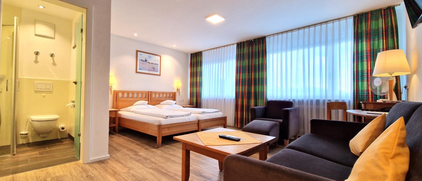 Spacious hotel room with a double bed, sofa, table, and bathroom. Light-colored curtains and colorful drapes adorn the windows., © TOMAS Spacious hotel room with a double bed, sofa, table, and bathroom. Light-colored curtains and colorful drapes adorn the windows., © TOMAS