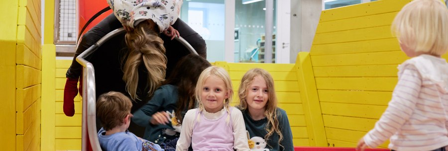 Children play on a slide in an indoor playground. The surroundings are colorful with yellow walls and play equipment., &copy; Julia Ochs