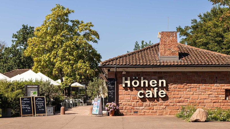 The H&ouml;hencaf&eacute; in Killesbergpark is a brick building with a terrace surrounded by trees and plants. White parasols are visible., &copy; Stuttgart-Marketing GmbH, Sarah Schmid