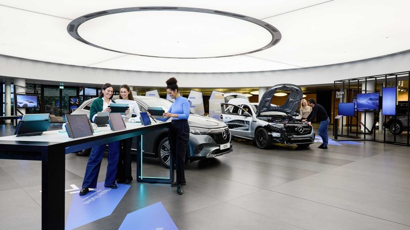 Moderner Ausstellungsraum mit Autos und interaktiven Stationen. Menschen nutzen Tablets, während andere ein Auto mit geöffneter Motorhaube betrachten., © Mercedes-Benz AG Moderner Ausstellungsraum mit Autos und interaktiven Stationen. Menschen nutzen Tablets, während andere ein Auto mit geöffneter Motorhaube betrachten., © Mercedes-Benz AG