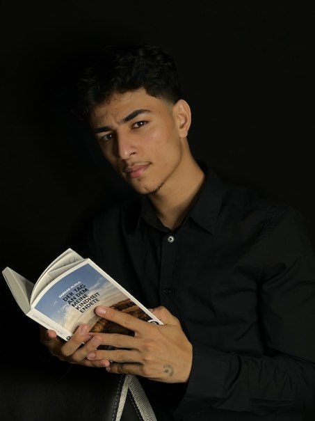 Ein Mann in schwarzem Hemd liest ein Buch mit dem Titel 'Der Tag, an dem meine Kindheit endete' vor schwarzem Hintergrund., © Theater Atelier Ein Mann in schwarzem Hemd liest ein Buch mit dem Titel 'Der Tag, an dem meine Kindheit endete' vor schwarzem Hintergrund., © Theater Atelier