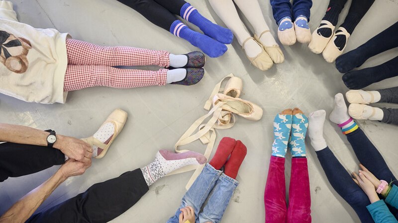Kinder und Erwachsene sitzen im Kreis auf dem Boden, ihre Beine ausgestreckt. Verschiedene bunte Socken und Ballettschuhe sind zu sehen., © Württembergische Staatstheater Stuttgart