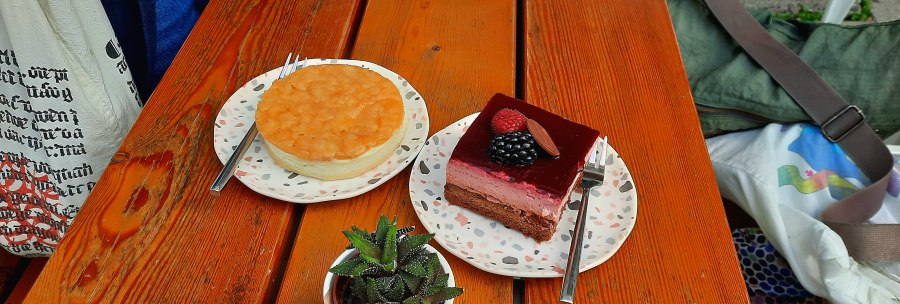 Two pieces of cake on plates with forks on a wooden table, next to a small plant., © SMG