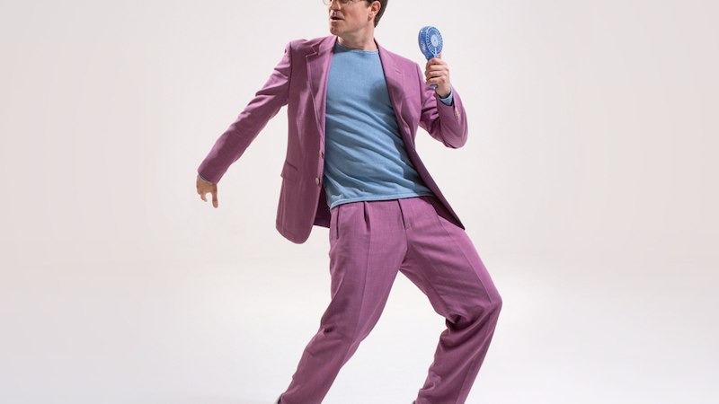 Person in rosa Anzug und blauer Bluse posiert mit einem kleinen Ventilator in der Hand auf hellem Hintergrund., © Renitenztheater Stuttgart e.V. Person in rosa Anzug und blauer Bluse posiert mit einem kleinen Ventilator in der Hand auf hellem Hintergrund., © Renitenztheater Stuttgart e.V.