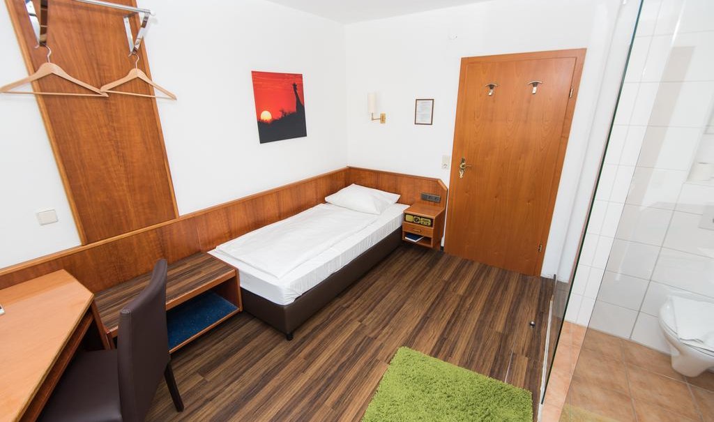Ein gemütliches Einzelzimmer mit Holzboden, Einzelbett, Schreibtisch und kleinem Badezimmer. An der Wand hängt ein Bild mit Sonnenuntergang., © ARNOTEL GmbH