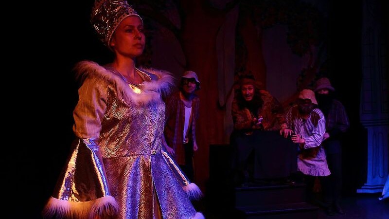 Eine Schauspielerin in einem prächtigen Kostüm der Schneekönigin steht auf einer Bühne, umgeben von weiteren Darstellern in einem dunklen, märchenhaften Setting., © Theater Atelier Eine Schauspielerin in einem prächtigen Kostüm der Schneekönigin steht auf einer Bühne, umgeben von weiteren Darstellern in einem dunklen, märchenhaften Setting., © Theater Atelier