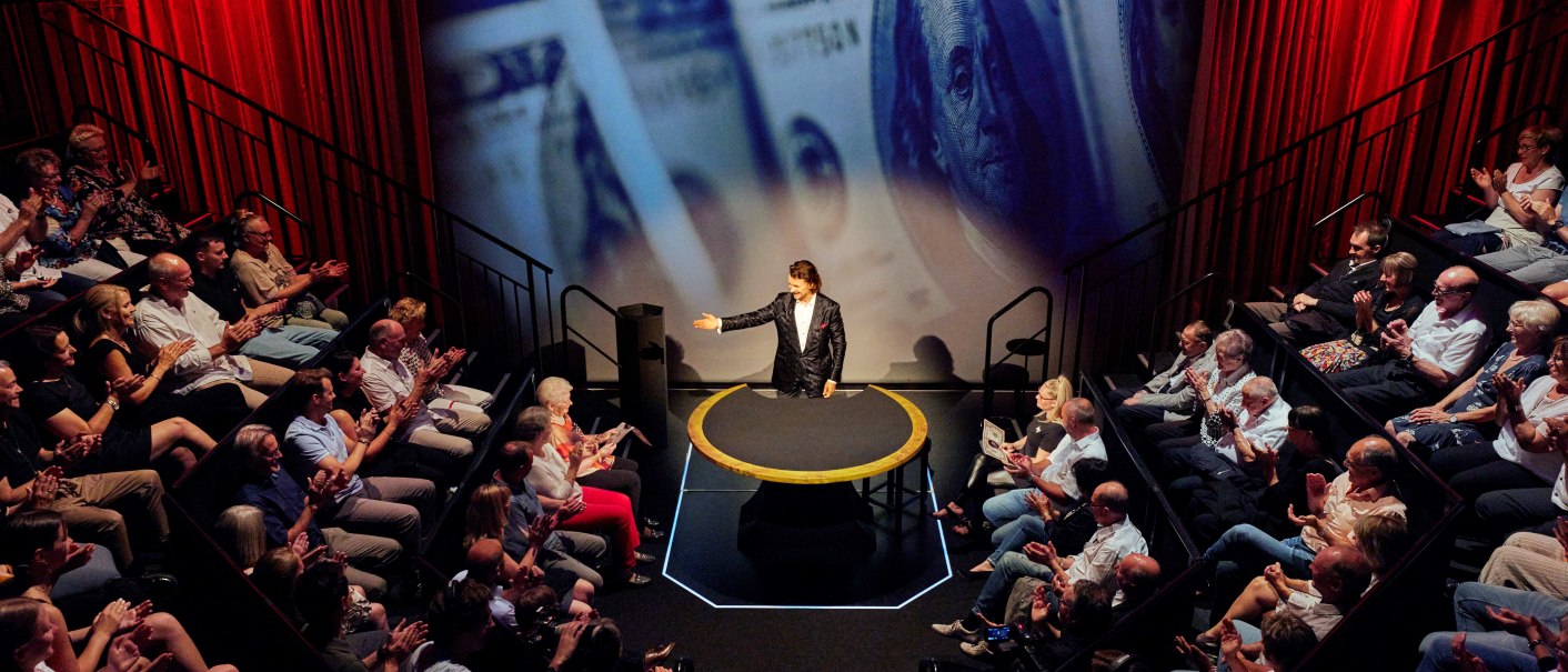A magician stands on a stage with a red curtain, surrounded by an applauding audience. Enlarged banknotes are projected in the background., © STROTMANNS Magic Lounge