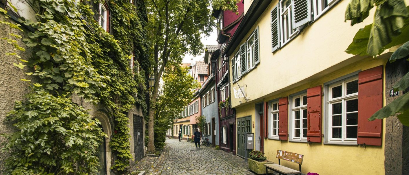 A charming, cobblestone alley in Esslingen, lined with colorful half-timbered houses and lush greenery. Two people are strolling down the street., © SMG Stuttgart Marketing GmbH - Sarah Schmid A charming, cobblestone alley in Esslingen, lined with colorful half-timbered houses and lush greenery. Two people are strolling down the street., © SMG Stuttgart Marketing GmbH - Sarah Schmid