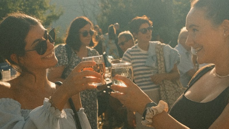 Two women are clinking wine glasses whilst standing in a cheerful crowd outdoors., © MyDegerloch Two women are clinking wine glasses whilst standing in a cheerful crowd outdoors., © MyDegerloch