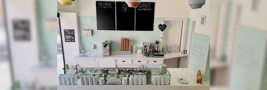 Ice cream parlor with counter, ice cream machine, coffee maker and ice cream cones. Pastel-colored tiles and lamps. Black boards with ice cream offers., © Zur Schleckerei Ice cream parlor with counter, ice cream machine, coffee maker and ice cream cones. Pastel-colored tiles and lamps. Black boards with ice cream offers., © Zur Schleckerei