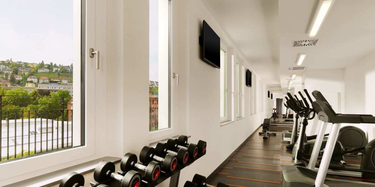 Modernes Fitnessstudio mit Laufbändern und Hanteln, helle Fenster mit Blick auf grüne Landschaft und Häuser im Hintergrund., © Aloft Stuttgart