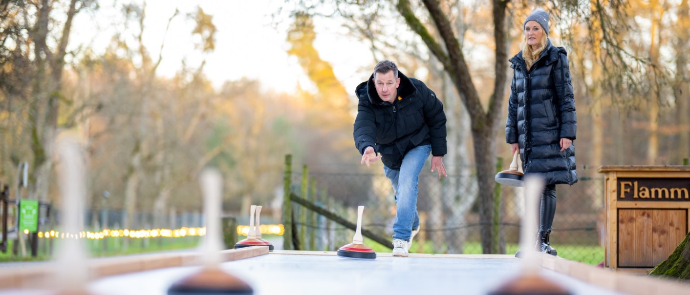 A man and a woman are playing curling outdoors. The man is just throwing a curling stone, while the woman watches. There are trees in the background., © Katzenbacher Hof A man and a woman are playing curling outdoors. The man is just throwing a curling stone, while the woman watches. There are trees in the background., © Katzenbacher Hof