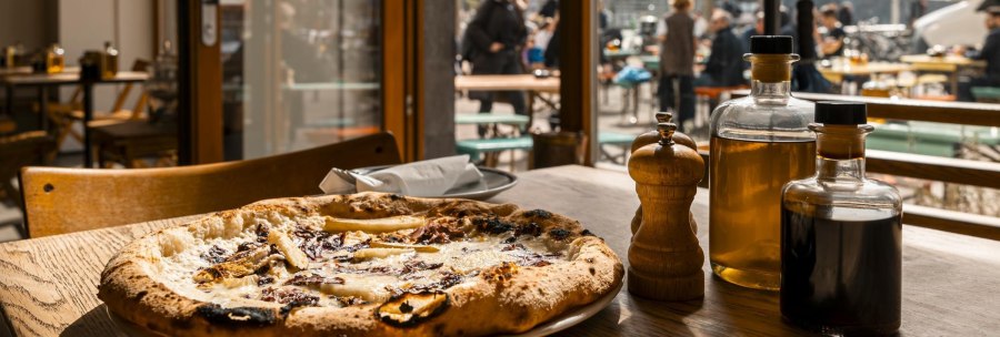 A pizza on a wooden table in a restaurant. People can be seen on a terrace in the background. There are bottles of oil and a pepper shaker on the table., © SMG Stuttgart Marketing GmbH - Sarah Schmid