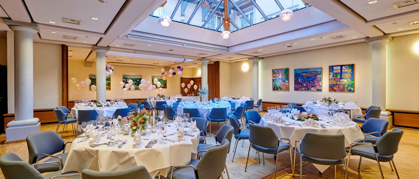 Elegant banquet hall with round tables, white tablecloths, flower arrangements and balloons. Large windowed ceiling and colorful paintings on the walls., © Hotel Gasthof Hasen Elegant banquet hall with round tables, white tablecloths, flower arrangements and balloons. Large windowed ceiling and colorful paintings on the walls., © Hotel Gasthof Hasen