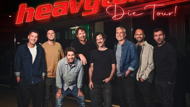 Eight men pose in front of a building with a glowing sign that reads "25 years of heavytones The Tour!". They smile into the camera., &copy; HEESEN KONZERTE GmbH