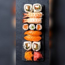 Eine Auswahl an Sushi, darunter Nigiri, Maki und Sashimi, kunstvoll auf einer schwarzen Platte angerichtet., © links im Bild Eine Auswahl an Sushi, darunter Nigiri, Maki und Sashimi, kunstvoll auf einer schwarzen Platte angerichtet., © links im Bild