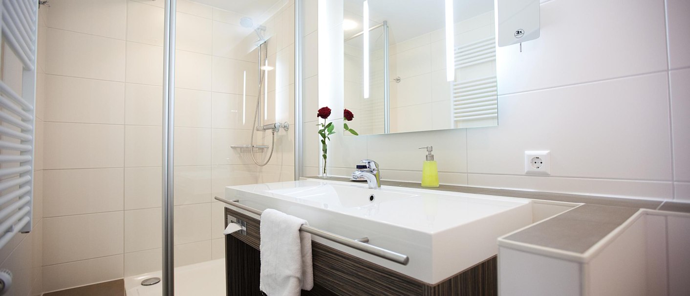 Modernes Badezimmer mit Glasdusche, großem Spiegel, Waschbecken und zwei roten Rosen. Handtücher und Seifenspender sind vorhanden., © Hotel Hirsch