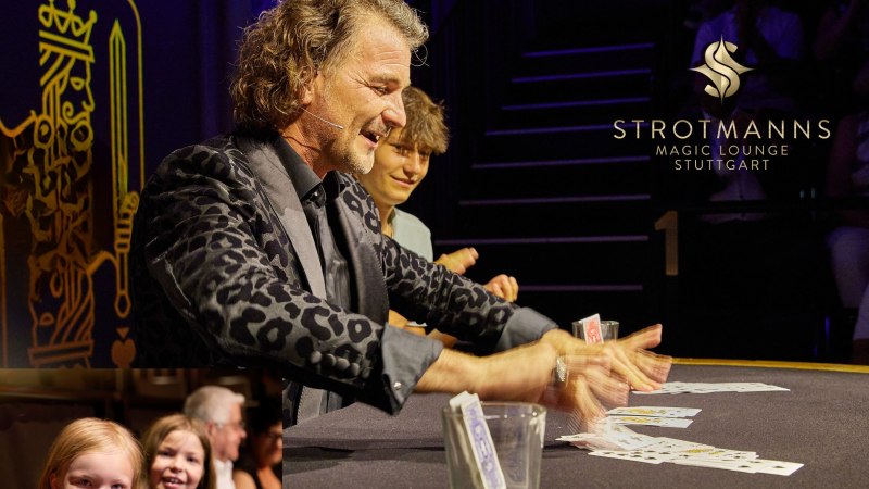 Ein Zauberkünstler führt Kartentricks in STROTMANNS Magic Lounge auf. Zuschauer, darunter Kinder, schauen fasziniert zu., © Strotmanns Magic Lounge Ein Zauberkünstler führt Kartentricks in STROTMANNS Magic Lounge auf. Zuschauer, darunter Kinder, schauen fasziniert zu., © Strotmanns Magic Lounge