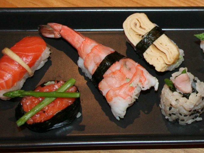 Ein schwarzer Teller mit verschiedenen Sushi-Varianten, darunter Lachs, Garnele, Omelett und Reisrollen, dekoriert mit Spargel und Algen., © Kultweine-Kochwelten-Kunstücke - Kochschule Sonja Lenz Ein schwarzer Teller mit verschiedenen Sushi-Varianten, darunter Lachs, Garnele, Omelett und Reisrollen, dekoriert mit Spargel und Algen., © Kultweine-Kochwelten-Kunstücke - Kochschule Sonja Lenz