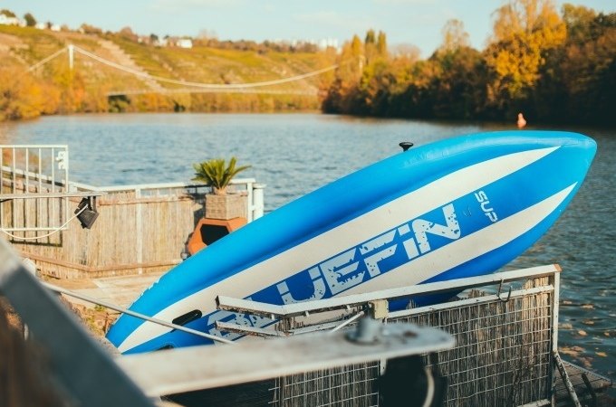 Ein Bluefin SUP-Board lehnt an einem Gel&auml;nder am Seeufer. Im Hintergrund ist eine H&auml;ngebr&uuml;cke und herbstliche Landschaft zu sehen., &copy; Cool-Tours StattReisen
