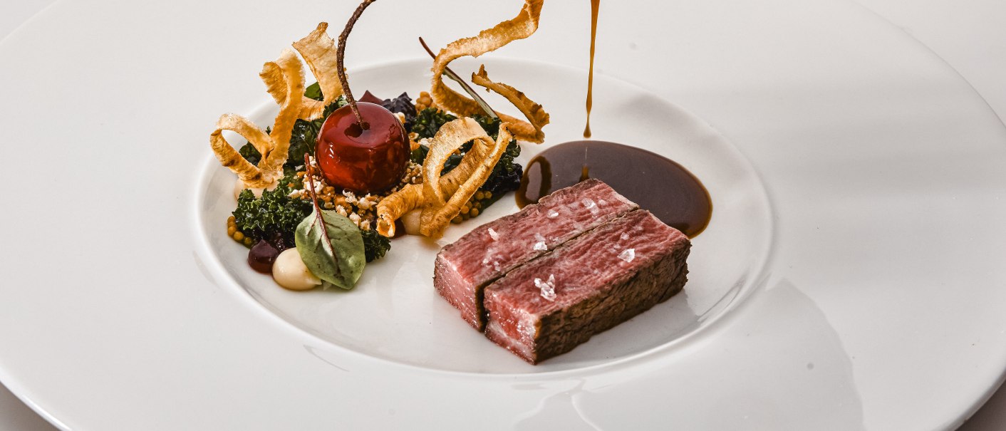 Ein kunstvoll angerichtetes Gourmetgericht mit Fleisch, Gemüse und einer dunklen Sauce, die aus einem Krug auf einen weißen Teller gegossen wird., © Zauberlehrling