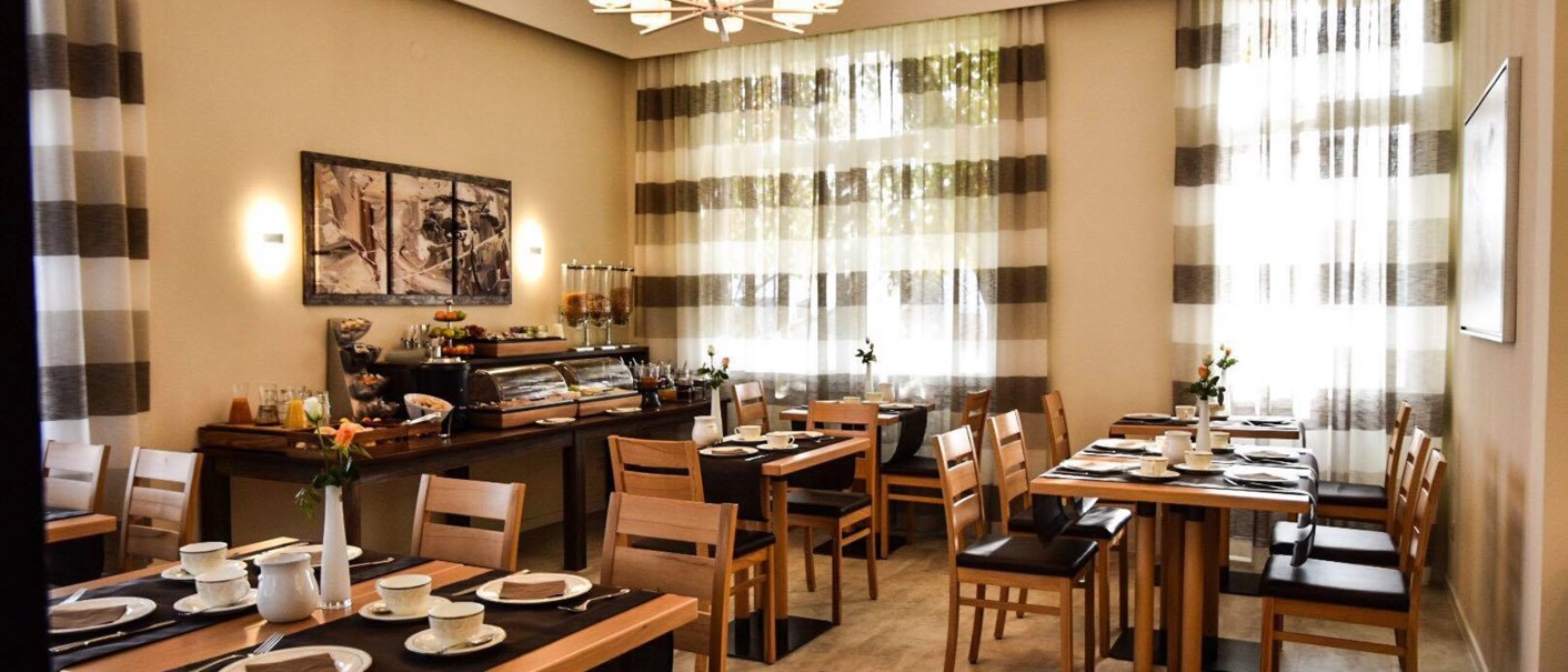 An elegant breakfast room with wooden furniture, laid tables and a buffet. Large windows with striped curtains let in light., &copy; TOMAS