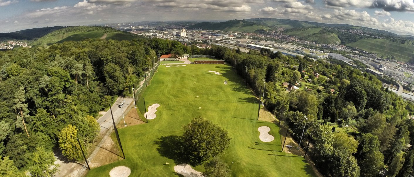 Aerial view of a golf course in Stuttgart, surrounded by trees and hills, with the city in the background under a cloudy sky., © GolfKultur Stuttgart Aerial view of a golf course in Stuttgart, surrounded by trees and hills, with the city in the background under a cloudy sky., © GolfKultur Stuttgart