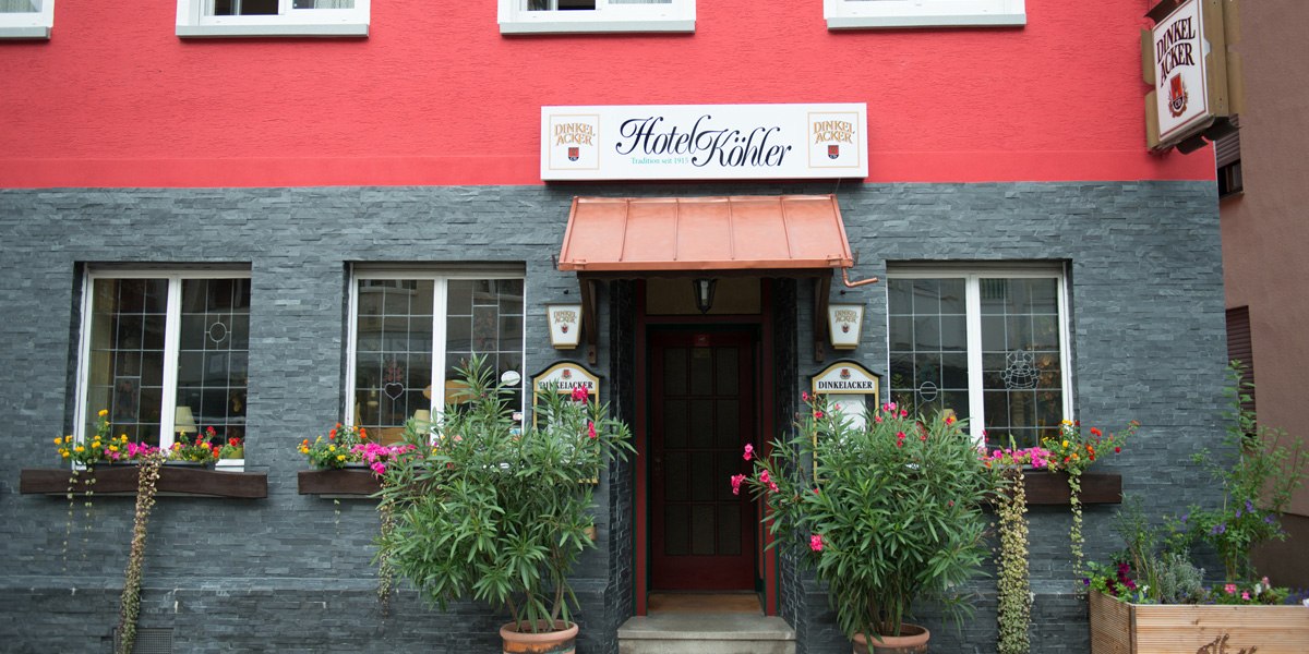 Das Hotel Köhler hat eine rote Fassade mit grauen Steinelementen. Vor dem Eingang stehen Pflanzen und bunte Blumen schmücken die Fenster., © Hotel Köhler