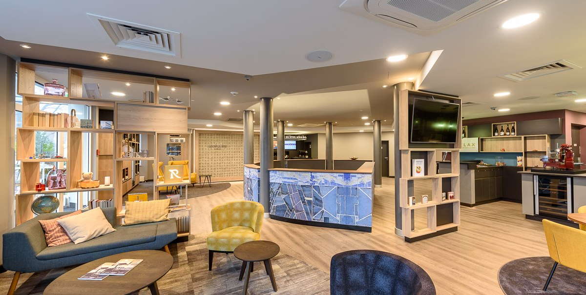 A modern hotel lobby with comfortable seating, shelves, and a front desk. Bright lighting and stylish furnishings dominate the space., &copy; TOMAS