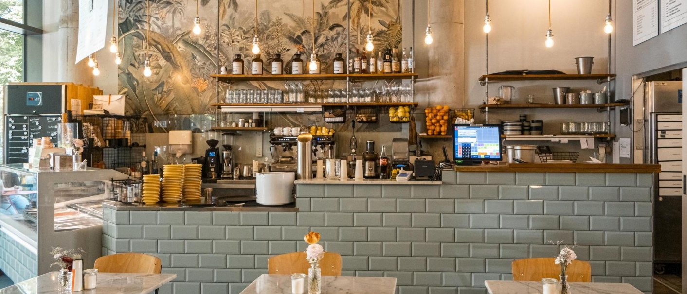 Ein gemütliches Café mit einer Theke, Regalen voller Gläser und Flaschen, und einer dekorativen Wand. Hängelampen beleuchten den Raum., © SMG, Sarah Schmid