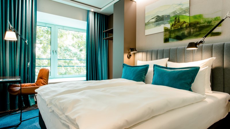 Modernes Hotelzimmer mit gro&szlig;em Bett, blauen Kissen und Vorh&auml;ngen. Ein Fenster bietet Blick ins Gr&uuml;ne. Ein Schreibtisch mit Stuhl steht daneben., &copy; Motel One - Stuttgart Hauptbahnhof