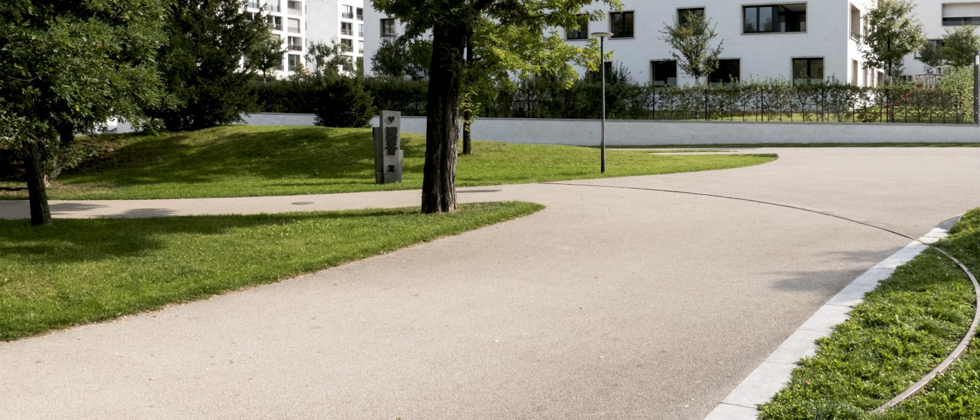 Ein gepflegter Park mit geschwungenen Gehwegen, Bäumen und modernen Wohngebäuden im Hintergrund. Ein Gedenkstein ist sichtbar., © Foto-Kunst Andreas Keller Ein gepflegter Park mit geschwungenen Gehwegen, Bäumen und modernen Wohngebäuden im Hintergrund. Ein Gedenkstein ist sichtbar., © Foto-Kunst Andreas Keller