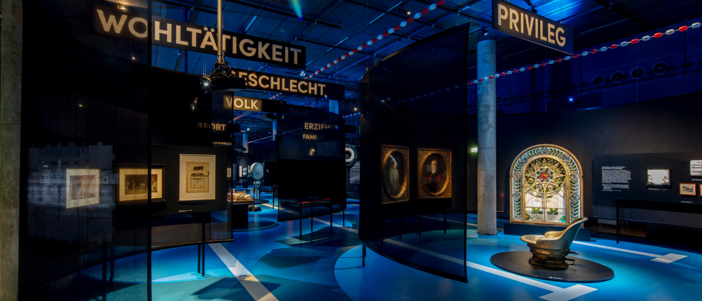 Museumsausstellung mit Kunstwerken, Schildern wie 'WOHLTÄTIGKEIT' und 'PRIVILEG', blauer Beleuchtung und einem dekorativen Fenster., © Frei_Schwimmen_Daniel_Stauch Museumsausstellung mit Kunstwerken, Schildern wie 'WOHLTÄTIGKEIT' und 'PRIVILEG', blauer Beleuchtung und einem dekorativen Fenster., © Frei_Schwimmen_Daniel_Stauch