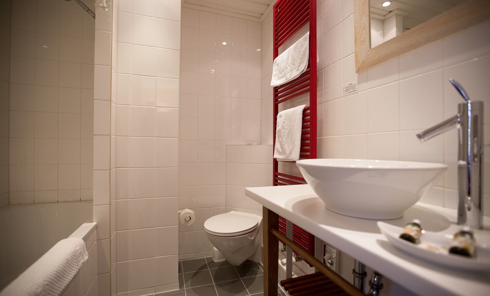 A modern bathroom with white ceramic tiles, a red towel warmer, and a large mirror. The walls are tiled, and the floor is gray., &copy; Hotel B&uuml;rkle