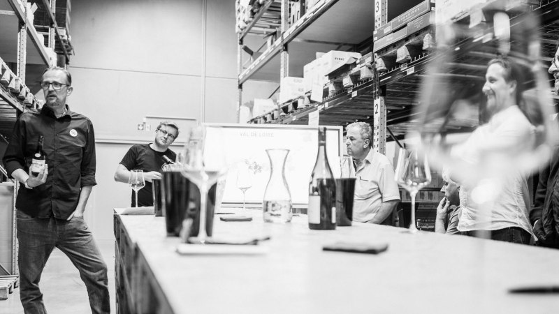 A group of people are standing in a storage room taking part in a wine tasting. Wine glasses and bottles can be seen in the foreground., © club traube
