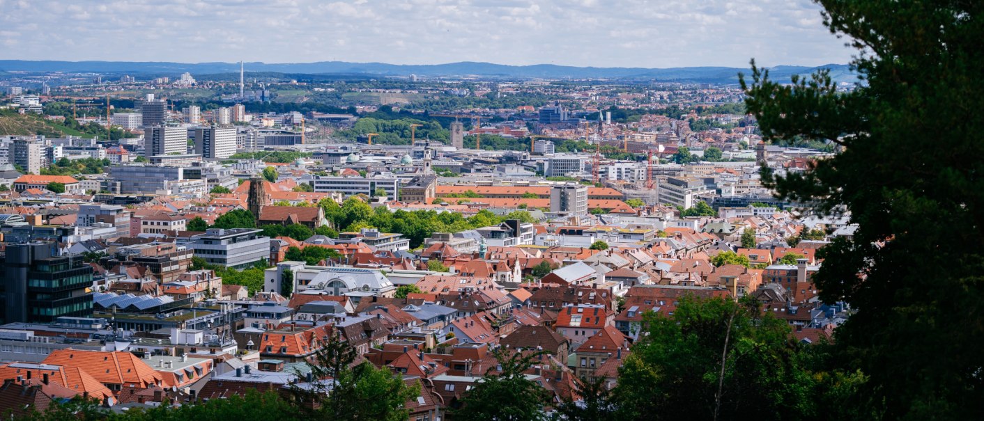 Panoramablick auf eine Stadt mit roten Dächern und modernen Gebäuden, umgeben von grünen Hügeln und bewölktem Himmel., © Thomas Niedermüller Panoramablick auf eine Stadt mit roten Dächern und modernen Gebäuden, umgeben von grünen Hügeln und bewölktem Himmel., © Thomas Niedermüller