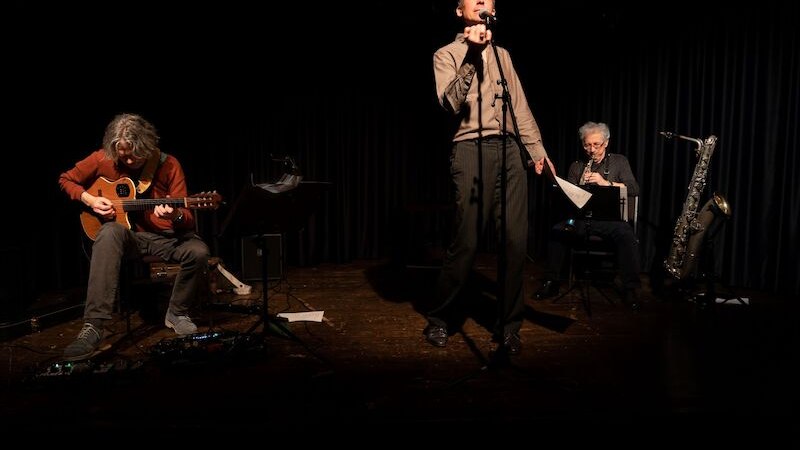 Drei Musiker auf einer Bühne: ein Gitarrist, ein Sänger am Mikrofon und ein Saxophonist. Die Bühne ist dunkel, der Fokus liegt auf den Künstlern., © Theaterhaus Stuttgart e.V.