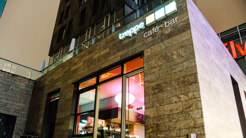 Modernes Café mit Glasfront, beleuchtetem Innenraum und Schriftzug "treppe café • bar" an der Fassade., © Café Treppe Modernes Café mit Glasfront, beleuchtetem Innenraum und Schriftzug "treppe café • bar" an der Fassade., © Café Treppe